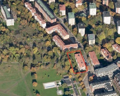 Flygfoto över ett stadsområde med en röd kontur som markerar en specifik byggnad bland bostadsstrukturer och grönområden.