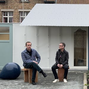 Två män som sitter utanför en byggnad med korrugerat plåttak, en på en träpall och den andra på en bänk, med ett blått sfäriskt föremål på marken bredvid sig.