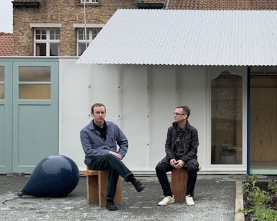 Två män som sitter utanför en byggnad med korrugerat plåttak, en på en träpall och den andra på en bänk, med ett blått sfäriskt föremål på marken bredvid sig.