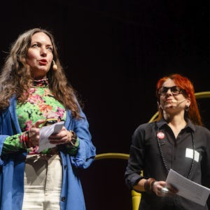 Två kvinnor presenterar på scenen, en talar med anteckningar i handen och den andra tittar på, med en suddig stadsbildsbakgrund på skärmen.