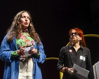 Två kvinnor presenterar på scenen, en talar med anteckningar i handen och den andra tittar på, med en suddig stadsbildsbakgrund på skärmen.