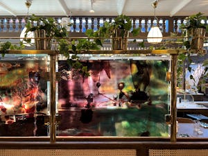 Konstnärliga glaspaneler med färgglada abstrakta mönster monterade på en guldram i en restaurang med hängande växter och takfönster.