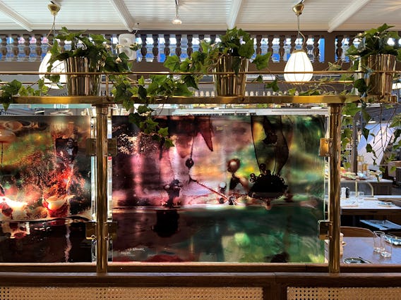Konstnärliga glaspaneler med färgglada abstrakta mönster monterade på en guldram i en restaurang med hängande växter och takfönster.