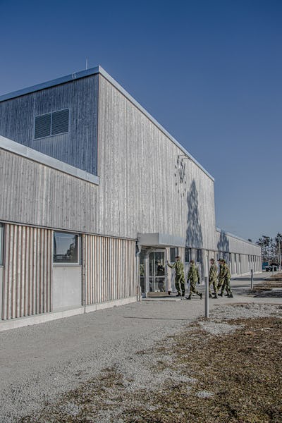 Modern betongbyggnad med vertikala träribbor, stora fönster och uniformerad personal som går utanför under en klarblå himmel.