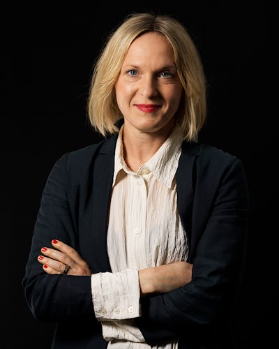 En kvinna med mellanlångt blont hår står mot en svart bakgrund, iförd en vit blus och svart kavaj, med armarna i kors och ler.