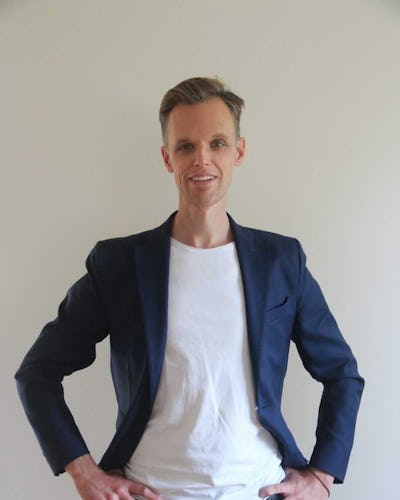 En person står mot en enkel bakgrund, klädd i en blå blazer över en vit skjorta och mörka byxor, tittade direkt in i kameran med händerna på höfterna.