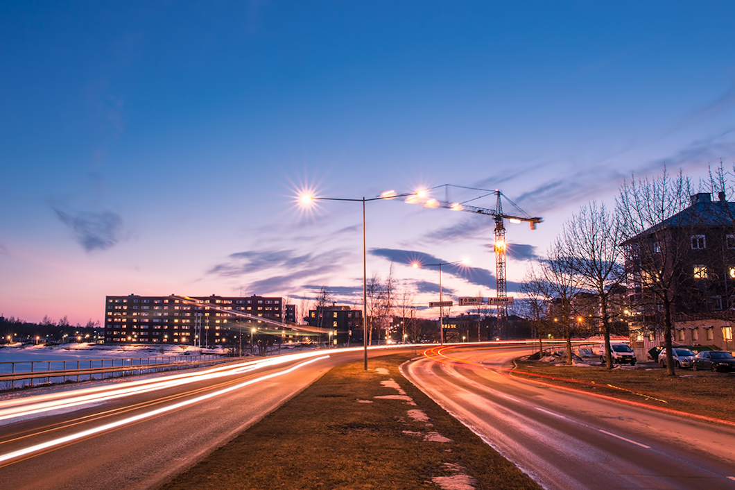 En stadsbild vid solnedgång med ljusspår från bilar på en väg, byggnader i bakgrunden och gatlyktor som lyser upp scenen.