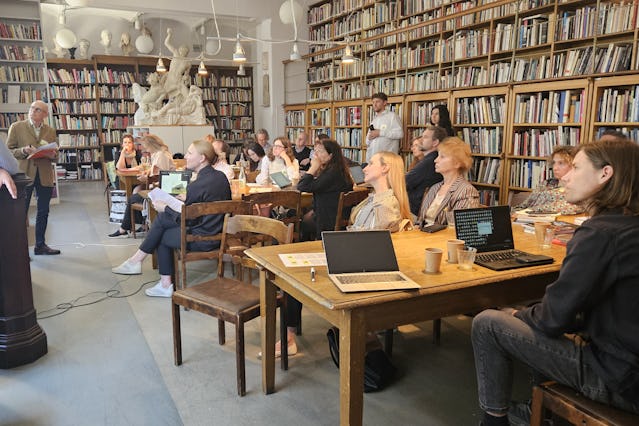 Personer sitter vid bord med bärbara datorer och böcker, och deltar i en föreläsning i ett bibliotek med bokhyllor och skulpturer i bakgrunden.