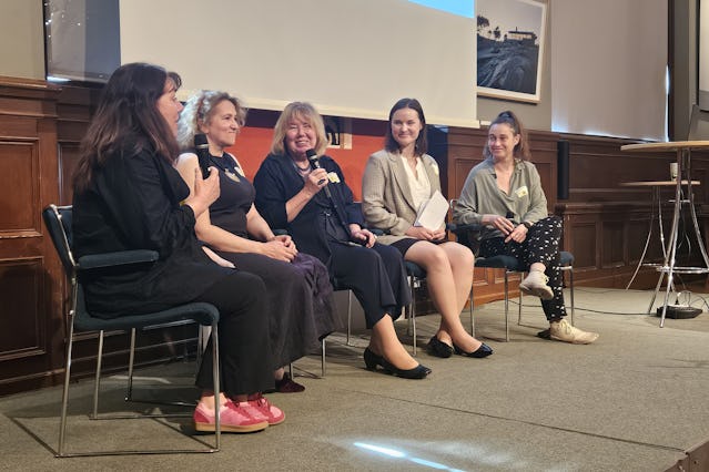 Fem kvinnor sitter på en scen, håller mikrofoner och deltar i en paneldiskussion. Bakom dem finns en vägg av trä och en projektorduk.
