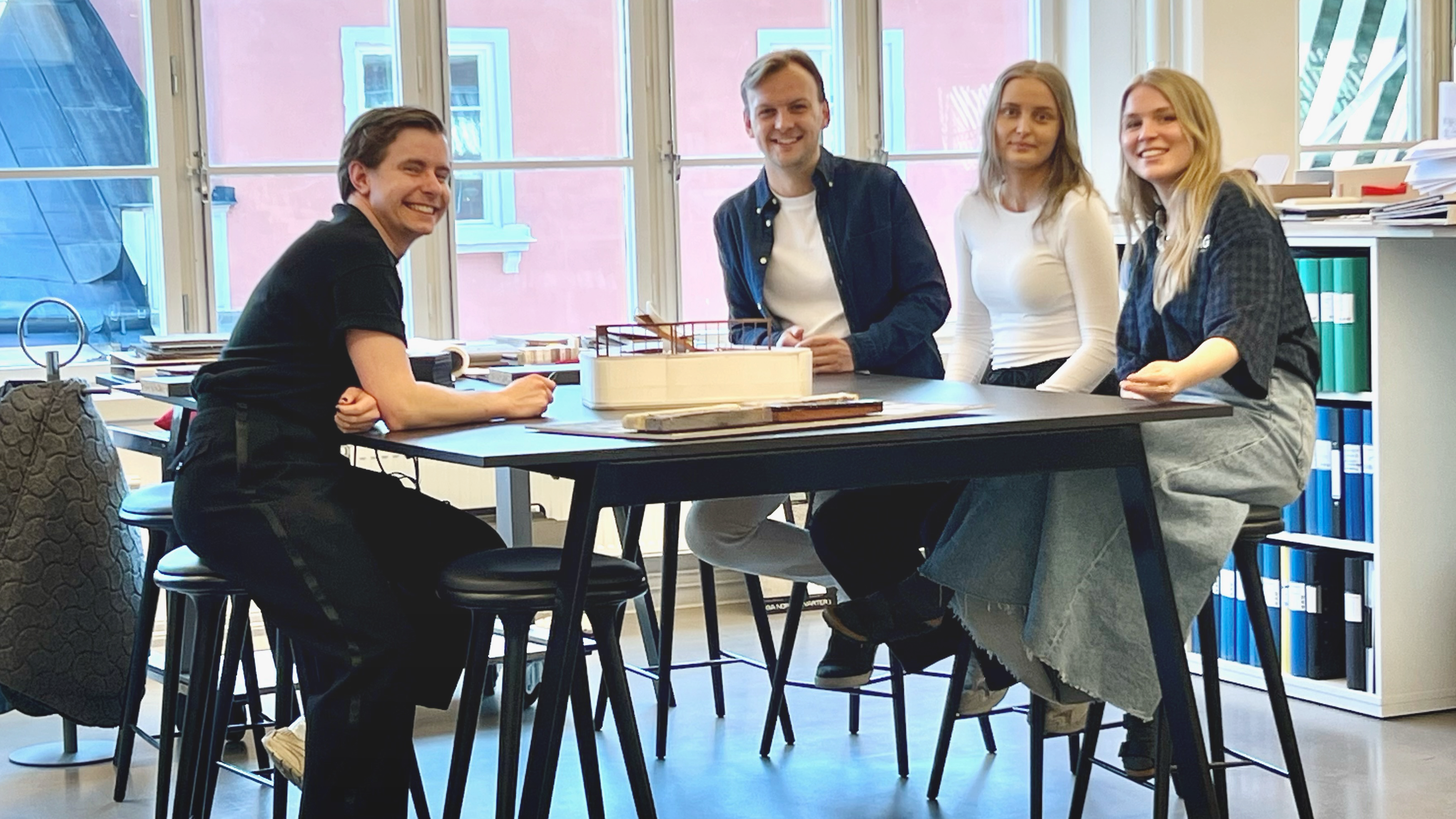 Fyra personer sitter runt ett bord i ett kontor och ler mot kameran. Det finns fönster i bakgrunden och kontorsmaterial på bordet.