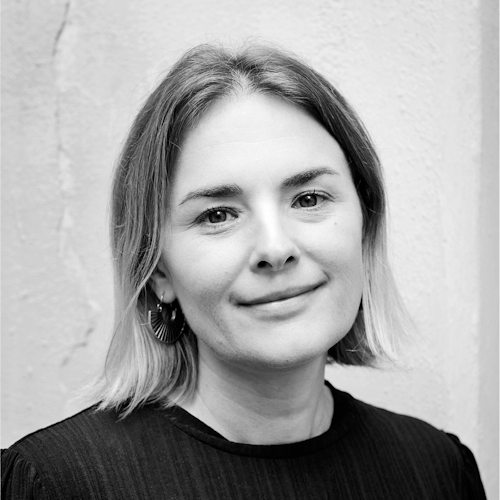 Svartvitt porträtt av en kvinna med axellångt hår, klädd i en mörk överdel och örhängen, som tittar rakt in i kameran med ett neutralt ansiktsuttryck. Bakgrunden består av en vägg med textur.