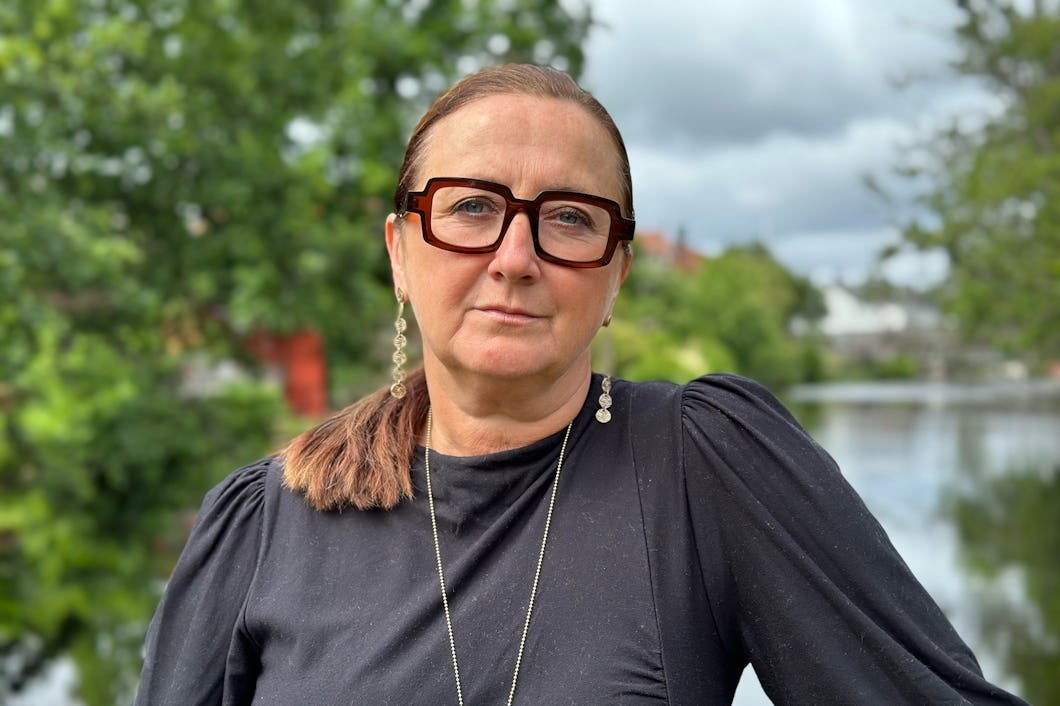 En person med glasögon och ett allvarligt ansiktsuttryck står utomhus framför vatten och växtlighet, klädd i en mörk överdel.
