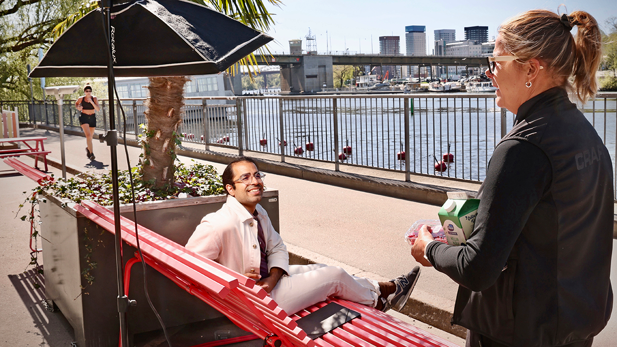 En man sitter på en bänk och ler mot en kvinna som står i närheten av vattnet. I bakgrunden finns en bro och byggnader.