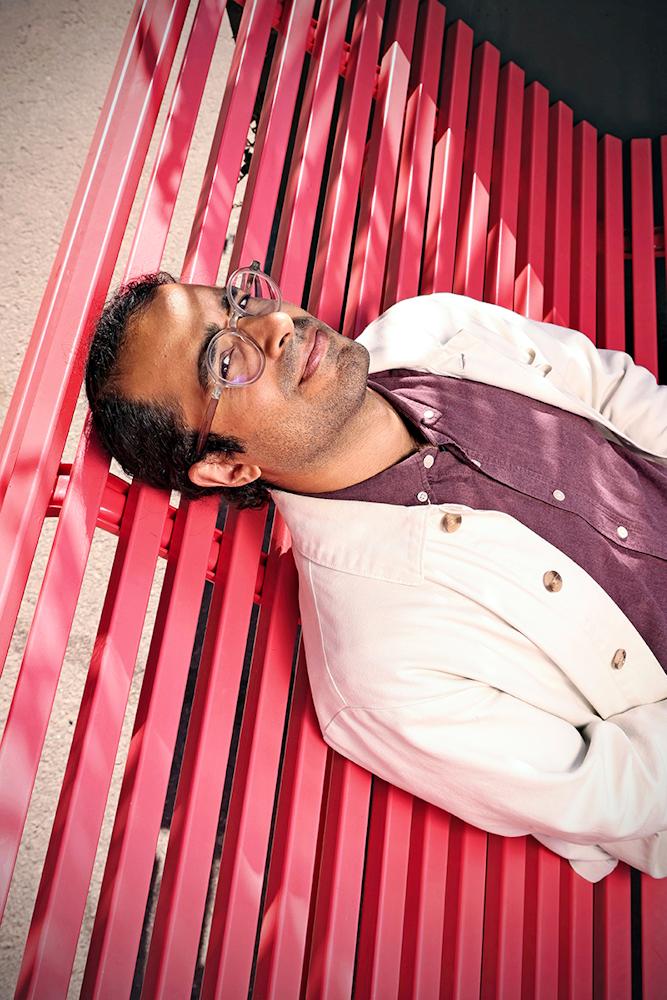 En person med glasögon, iklädd en jacka och en mörkröd skjorta, ligger på en röd bänk med spjälor och tittar uppåt.