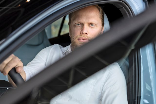 En person i vit skjorta sitter i förarsätet på en bil, med en hand på ratten och tittar genom den öppna sidorutan.