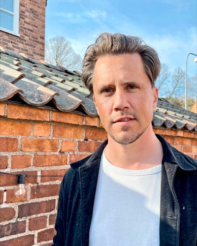 En man med kort, fixat hår och ljus skäggväxt står framför en tegelvägg. Han har på sig en vit skjorta och en mörk jacka. I bakgrunden syns träd och delar av ett hustak.