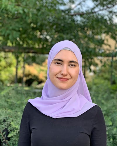 En kvinna med hijab och en svart skjorta står utomhus med växter i bakgrunden.