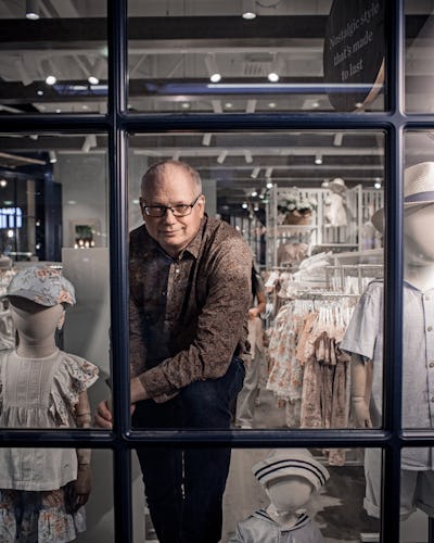En man med glasögon tittar genom ett butiksskyltfönster som visar barnkläder och skyltdockor med hattar och klänningar. Butikens inre syns i bakgrunden.