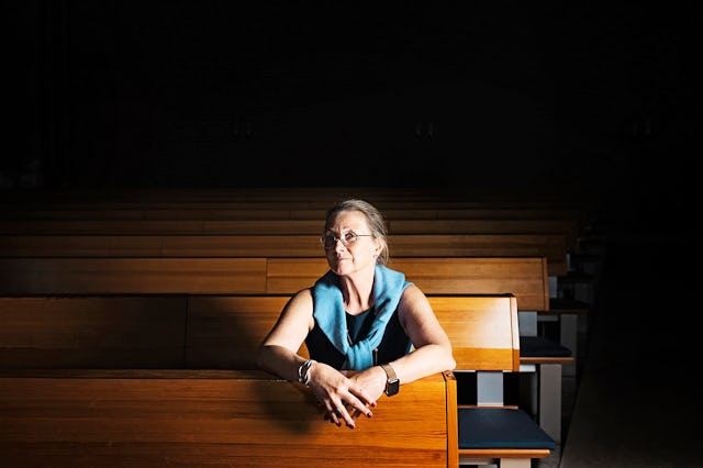 En kvinna med glasögon och en blå sjal sitter ensam på en bänk i kyrkan, där ljuset är svagt.