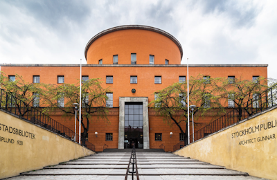 Stockholms stadsbibliotek av arkitekten Gunnar Asplund, en stor orange byggnad färdigställd 1928, sedd framifrån med träd på båda sidor. Utsikten inramas av trappor som leder upp.