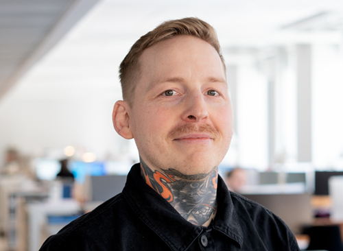 En man med mustasch och tatueringar på halsen står i en kontorsmiljö, iklädd en svart skjorta.