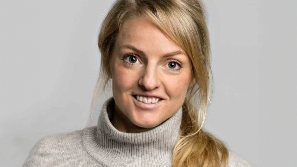 Kvinna med blont hår i en låg hästsvans, klädd i en ljus polotröja, ler mot kameran. Bakgrunden är enkel.