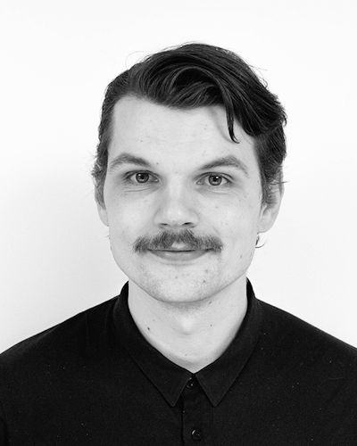 Svartvitt porträtt av en man med mustasch och kort hår, iförd en mörk skjorta med krage. Han tittar rakt in i kameran med ett neutralt ansiktsuttryck.