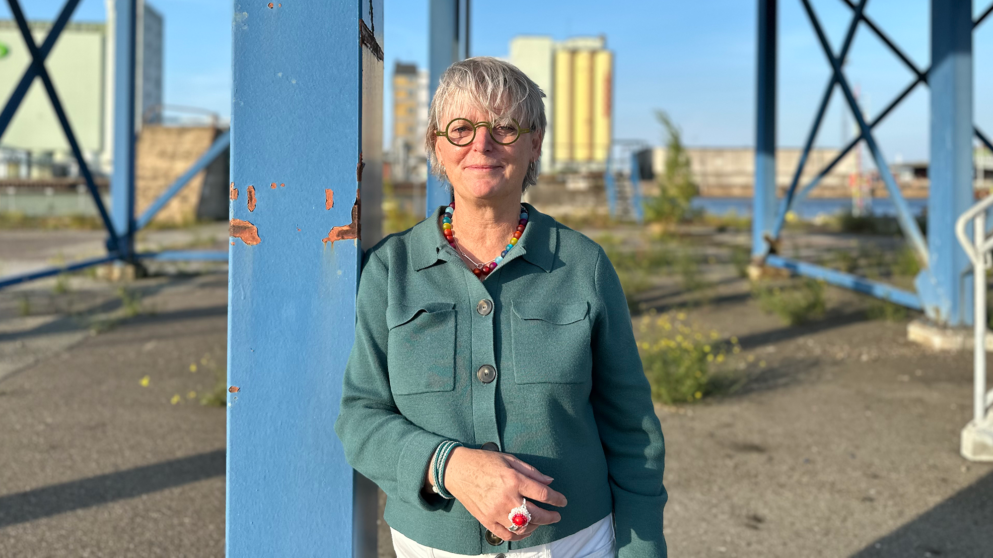 En kvinna med kort grått hår och glasögon står utomhus intill en blå metallstruktur på en soldag. Personen har en teal jacka och vita byxor på sig.