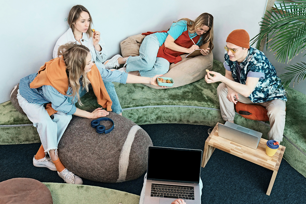 En grupp på fyra personer sitter på stolar och kuddar i en avslappnad miljö med bärbara datorer och tilltugg. En person sträcker sig för att ge något till en annan.