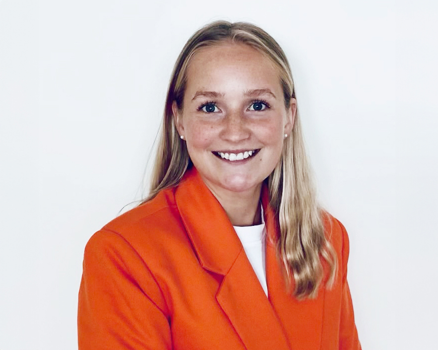 En kvinna med långt blont hår, iklädd en orange kavaj och en vit topp, ler framför en vit bakgrund.