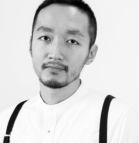 Svartvitt porträtt av en man med kort hår och skägg, klädd i vit skjorta och hängslen, tittar rakt in i kameran.