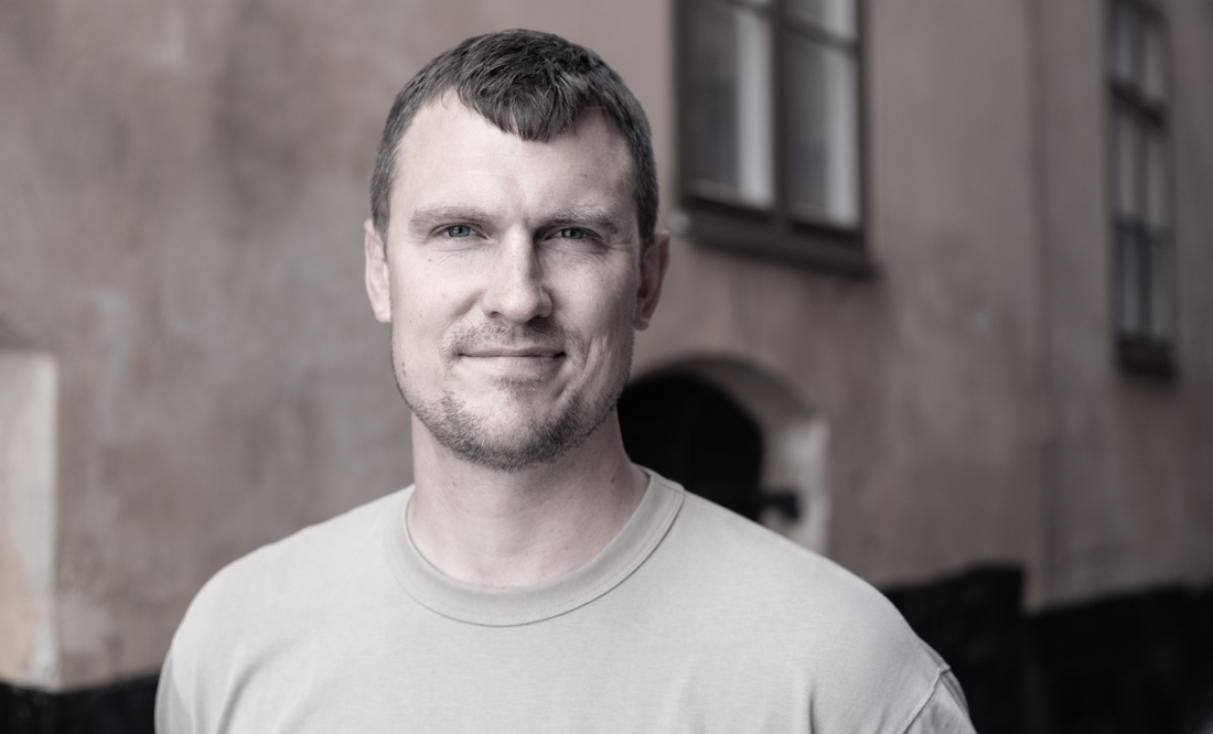 En man i t-shirt ler utomhus med en oskarp byggnad i bakgrunden.