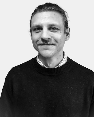En man med mustasch, klädd i en randig skjorta och mörk tröja, ler mot en enkel bakgrund.