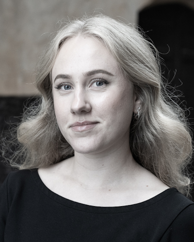 En kvinna med ljust, vågigt hår har på sig en svart överdel och ler svagt, stående mot en oskarp bakgrund.