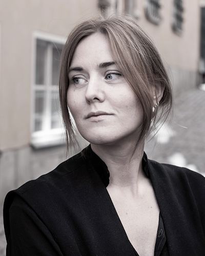 En kvinna står utomhus, klädd i en mörk överdel och tittar åt sidan med ett neutralt ansiktsuttryck. I bakgrunden syns en smal gata och byggnader.