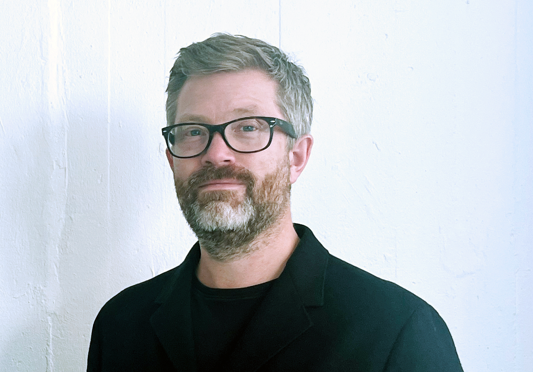 En man med glasögon och skägg står mot en vit vägg, iklädd en svart jacka och skjorta.