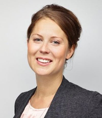 En kvinna med brunt hår ler mot kameran. Hon har en grå blazer över en ljus blus och står framför en enkel bakgrund.