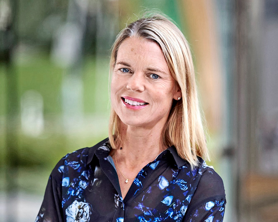 En kvinna med axellångt blont hår ler och har på sig en mörk blus med blommönster. Bakgrunden är oskarp grönska.