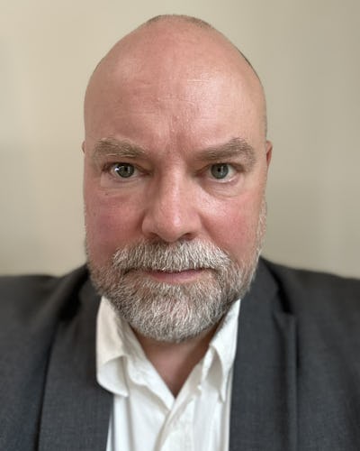 En skäggig person i mörk kavaj och vit skjorta tittar rakt in i kameran.