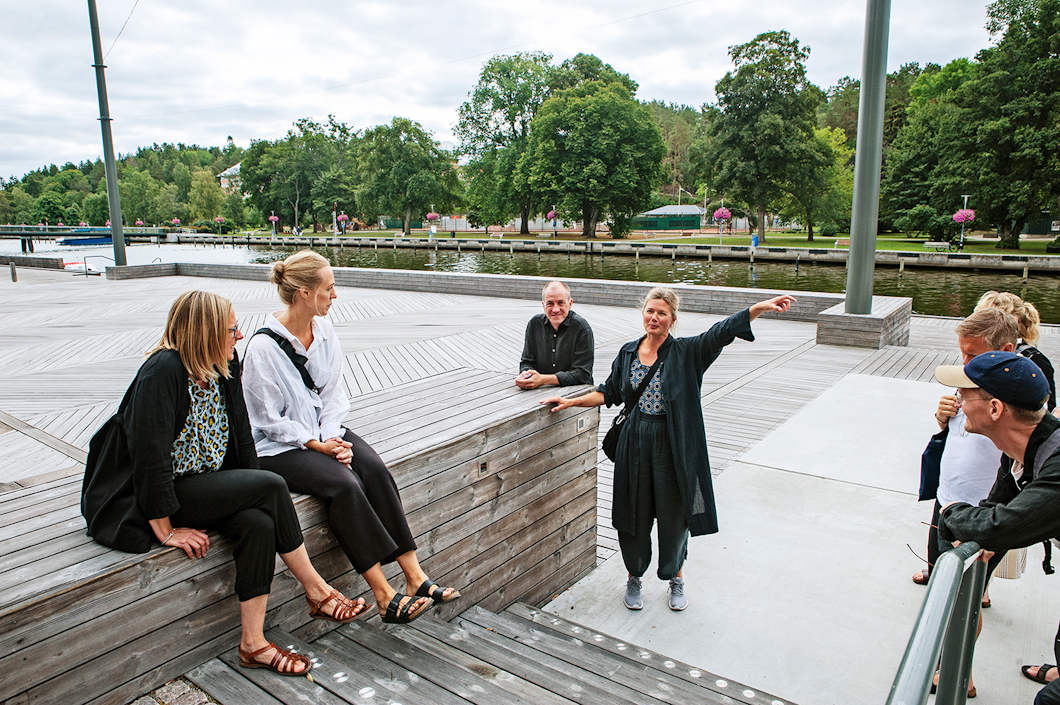 En grupp personer samlades på trätrappor nära en flod, med en person som pekade mot något. I bakgrunden finns träd och en molnig himmel.