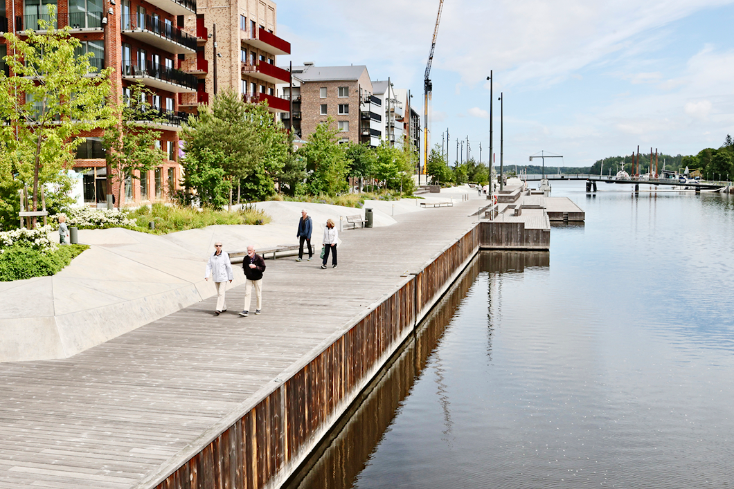 Människor går längs en träbrygga bredvid en flod, omgivna av moderna byggnader och växtlighet under en delvis molnig himmel.