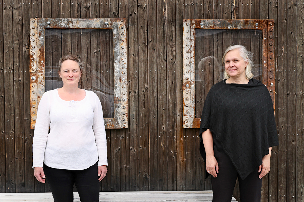 Två kvinnor står framför en trävägg med två rostiga, inramade öppningar. Den ena har en vit skjorta, den andra en svart poncho.