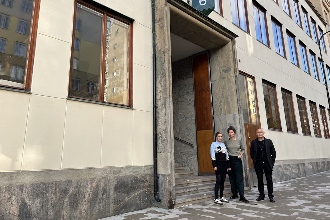 Tre personer står utanför en byggnadsingång med en skylt med siffran 6. Byggnaden har stora fönster och en fasad av sten.