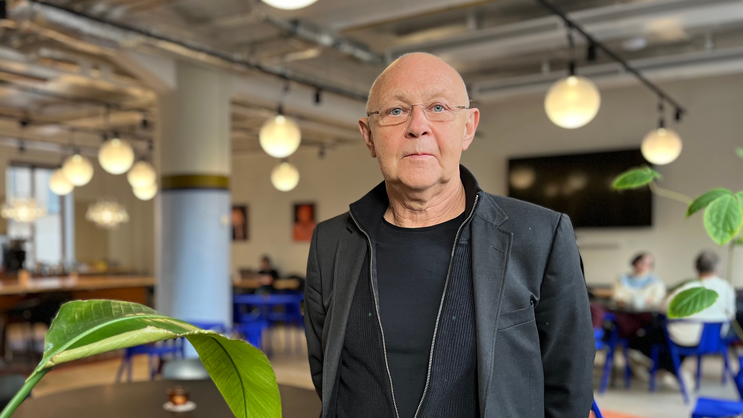 En man med glasögon och svart jacka står inomhus, med växter och människor i bakgrunden.
