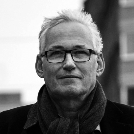 En svartvit bild av en man med kort hår och glasögon, iförd halsduk och rock, som tittar rakt in i kameran.