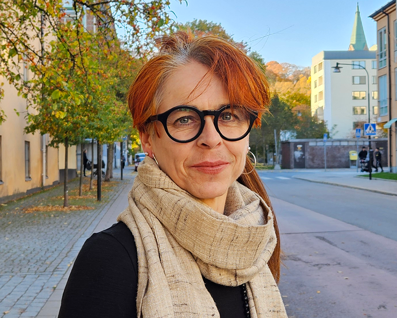 En person med rött hår och glasögon står på en gata med byggnader och träd i höstskrud, klädd i en svart topp och beige halsduk.