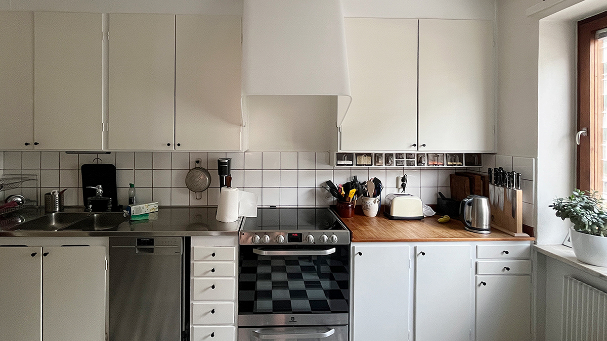 Ett kök med vita skåp, en diskbänk i rostfritt stål, en elektrisk spis med fläkt, en bänkskiva av trä, små apparater och ett fönster med en krukväxt.