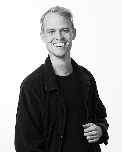 En person med kort, ljust hår ler och har på sig en mörk jacka över en mörk skjorta, stående mot en enkel bakgrund.