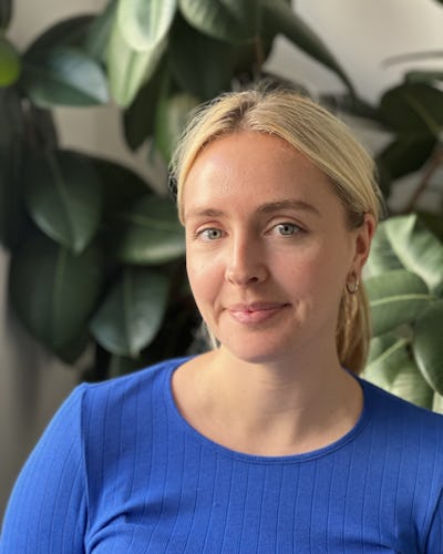 En person med ljust hår i en blå tröja står framför gröna växter.