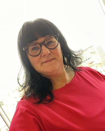 En kvinna med mörkt hår och glasögon har på sig en rosa topp. Hon står framför ett fönster med en stad i bakgrunden.